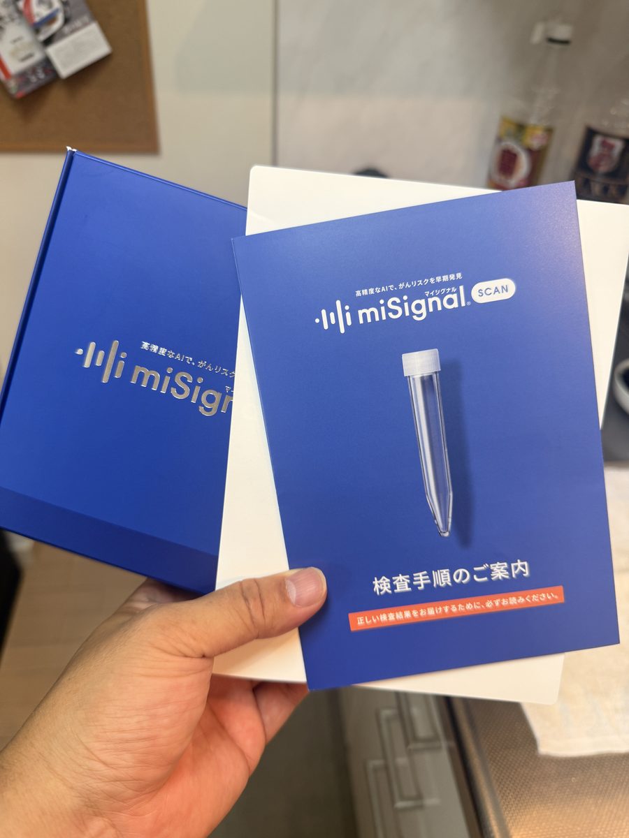 miSignalの検査キットの青い箱と、検査手順の案内カードを手に持っている